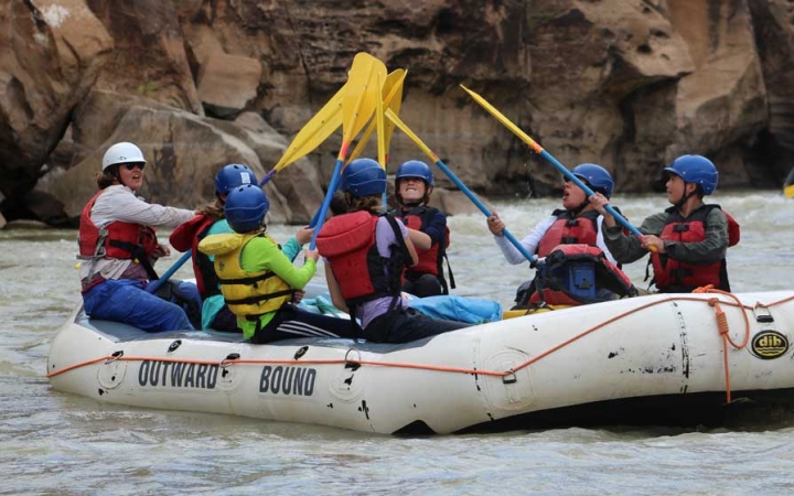 A group of students wearing safety gear sit in a raft and raise their paddles into the air.